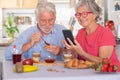 Beautiful elderly couple having breakfast at home, woman using smart phone. Senior people relaxed and happy Royalty Free Stock Photo