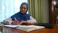 Beautiful confident Arab Muslim woman in blue hijab, makes notes on notepad while telecommute on laptop from home office