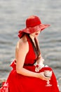 A beautiful elegant lady in a red hat and a long red dress on the beach with a glass of wine in her hands. Summer holidays Royalty Free Stock Photo