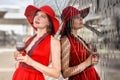 A beautiful elegant lady in a red hat and a long red dress on the embankment by the mirrored wall with a glass of wine in her Royalty Free Stock Photo