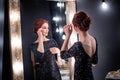 Beautiful, elegant woman in black evening dress stands next to dark high mirror