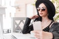 Beautiful elegant woman in sunglasses in dress in leather jacket with hat sits at table in cafe and makes selfie on mobile phone. Royalty Free Stock Photo