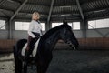 Beautiful elegant young blonde girl sits on a her black horse dressing uniform competition white blouse shirt and brown pants. Royalty Free Stock Photo