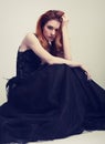Beautiful elegant young model with bright redhead hairstyle sitting in fashion chic black dress with long skirt on studio