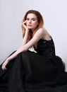 Beautiful elegant young model with bright redhead hairstyle sitting in fashion chic black dress with long skirt on studio grey