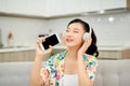 Beautiful emotional pleased young woman on sofa at home listening music with headphones holding mobile phone singing Royalty Free Stock Photo