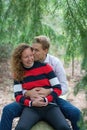 Beautiful enamored young couple sitting on a tree trunk in the forest. Facial expression is in love, man kisses the woman