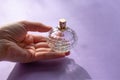 Beautiful faceted glassy tranclucent bottle of eau de parfum toilet water on purple background in woman hand.