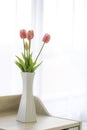 Beautiful fake pink tulips in white vase on wooden table in bed room with soft sun light and white curtains background