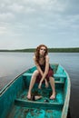 Beautiful fashion redhead woman sit in boat. Beauty romantic portrait girl in red vinous dress in wooden boat on lake