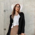 Beautiful fashion young woman in vintage white hat in trendy black blazer in sexy top in elegant glasses stands near vintage wall Royalty Free Stock Photo