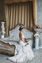 Beautiful and fashionable brunette model girl in stylish wedding dress with naked shoulders sits on the bed with bouquet Royalty Free Stock Photo