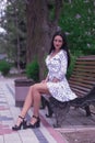 beautiful fashionable brunette woman adult sitting on a bench the park on the plants backgraund in white dress with polka dots Royalty Free Stock Photo