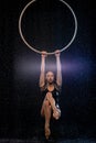 Beautiful female acrobat hanging on aerial hoop under rain on black background
