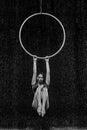 Beautiful female acrobat hanging on aerial hoop under rain on black background