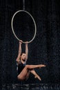 Beautiful female acrobat hanging with graceful pose on aerial hoop under rain on black background