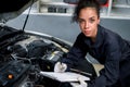 Beautiful female auto mechanic working in garage, car service technician woman holding file paper and checking car engine, Royalty Free Stock Photo