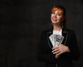 Beautiful female in black jacket and white shirt looking at camera with charming smile holding plenty of dollars in hands