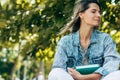 A beautiful female with closed eyes with a book in hand while sitting on the bench on a sunny day in the street. A pretty woman in