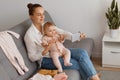 Beautiful female with dark hair and bun hairstyle wearing casual clothing sitting on cough with toddler baby with nipple in hand, Royalty Free Stock Photo