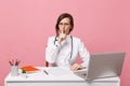 Beautiful female doctor sits at desk works on computer with medical document in hospital isolated on pastel pink wall