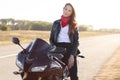 Beautiful female driver sits on black fast motobike, dressed leather jacket and red bandana, travels around country by motobike,