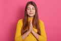 Beautiful female with long hair praying with closed eyes, keeping palm together, posing isolated over pink background, wearing