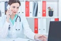 Beautiful Caucasian female medicine doctor working at the laptop and talking by mobile phone. Royalty Free Stock Photo