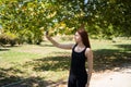 Beautiful female smiling and taking selfie via smartphone while standing on sunlit park alley