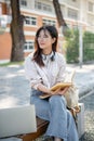 A beautiful female student is sitting on a bench in a campus garden with her laptop and a book Royalty Free Stock Photo