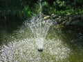 Beautiful fountain in garden pond against background of emerald green of shady summer garden. Freshness of water jets creates a mo Royalty Free Stock Photo