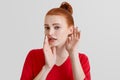 Beautiful foxy woman tries to overhear or eavesdrop someonen`s gossiping, keeps hand near ear, looks with secrecy, isolated over w Royalty Free Stock Photo