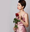 Beautiful fragile and tender brunette woman with short hair in pink silk dress and earrings with red rose in her arms Royalty Free Stock Photo