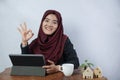 A beautiful, friendly young Muslim business woman smile and thumbs up okay at camera in hijab dress and suit using laptop