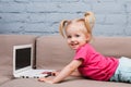 Beautiful funny blonde girl a child of two years is lying on the couch indoors and uses a white laptop computer technology with co Royalty Free Stock Photo