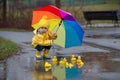 Beautiful funny blonde toddler boy with rubber ducks and colorful umbrella, jumping in puddles and playing in the rain Royalty Free Stock Photo