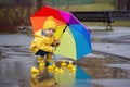 Beautiful funny blonde toddler boy with rubber ducks and colorful umbrella, jumping in puddles and playing in the rain Royalty Free Stock Photo