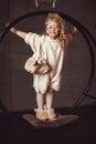 A beautiful and funny curly blonde in a white fluffy sweater stands on a swing against a dark background, smiles broadly Royalty Free Stock Photo