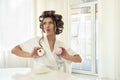 Beautiful funny young brunette woman in hair curlers holding two cupcakes in both hands near her breast sitting with cup of coffee Royalty Free Stock Photo