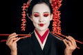 Beautiful geisha in black kimono with red flowers in hair and closed eyes holding chopsticks isolated on black Royalty Free Stock Photo