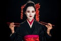 Beautiful geisha in black kimono with red flowers in hair holding chopsticks isolated on black Royalty Free Stock Photo