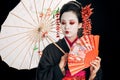 Geisha in black kimono with red flowers in hair holding traditional asian umbrella and hand fan isolated on black Royalty Free Stock Photo