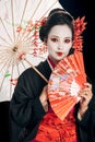 Beautiful geisha in black kimono with red flowers in hair holding traditional asian umbrella and hand fan isolated on Royalty Free Stock Photo