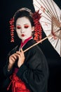 Geisha in black kimono with red flowers in hair holding traditional asian umbrella isolated on black Royalty Free Stock Photo