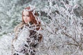 Beautiful gentle girl with red hair in a fur vest standing in a snowy forest with iniem on the branches of trees