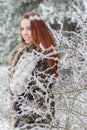 Beautiful gentle girl with red hair in a fur vest standing in a snowy forest with iniem on the branches of trees