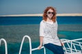 Beautiful ginger woman in sun glasses sits on a white yacht in a sea with clear turquoise water. Relaxation at summer vacation Royalty Free Stock Photo