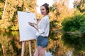 Beautiful girl artist, summer park by lake, river pond, draws picture, palette with paints, brush canvas painting, easel Royalty Free Stock Photo