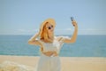 A beautiful girl on the background of the beach, sea and ocean makes a photo on the phone. Blonde in white outfit on vacation