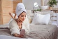 Beautiful girl in bathrobe and with towel on her head making selfie using smartphone on the bed at cozy hotel room. Royalty Free Stock Photo